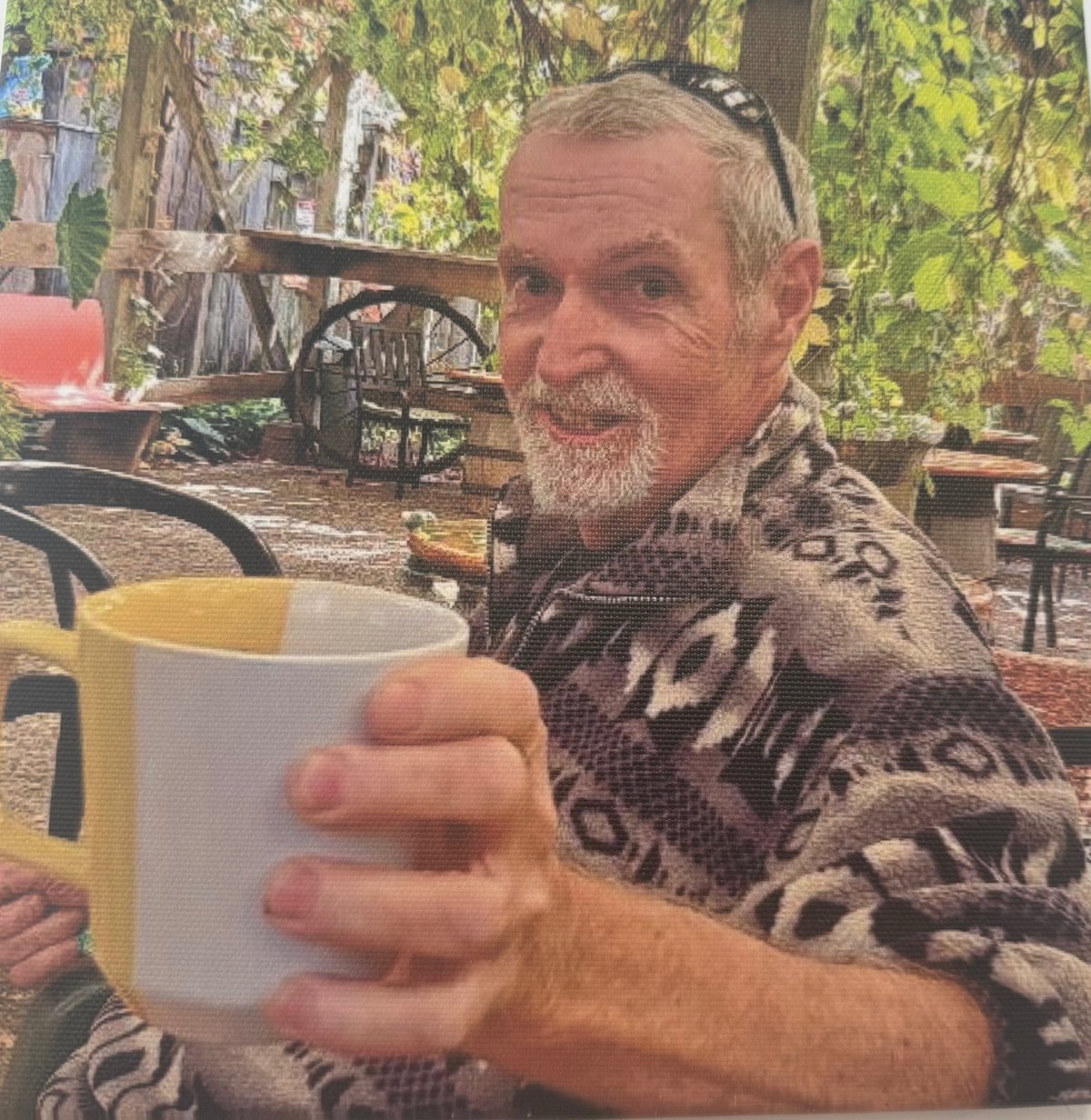 Middle-aged man with beard holding a white mug in an outdoor garden setting with trees and furniture visible in the background
