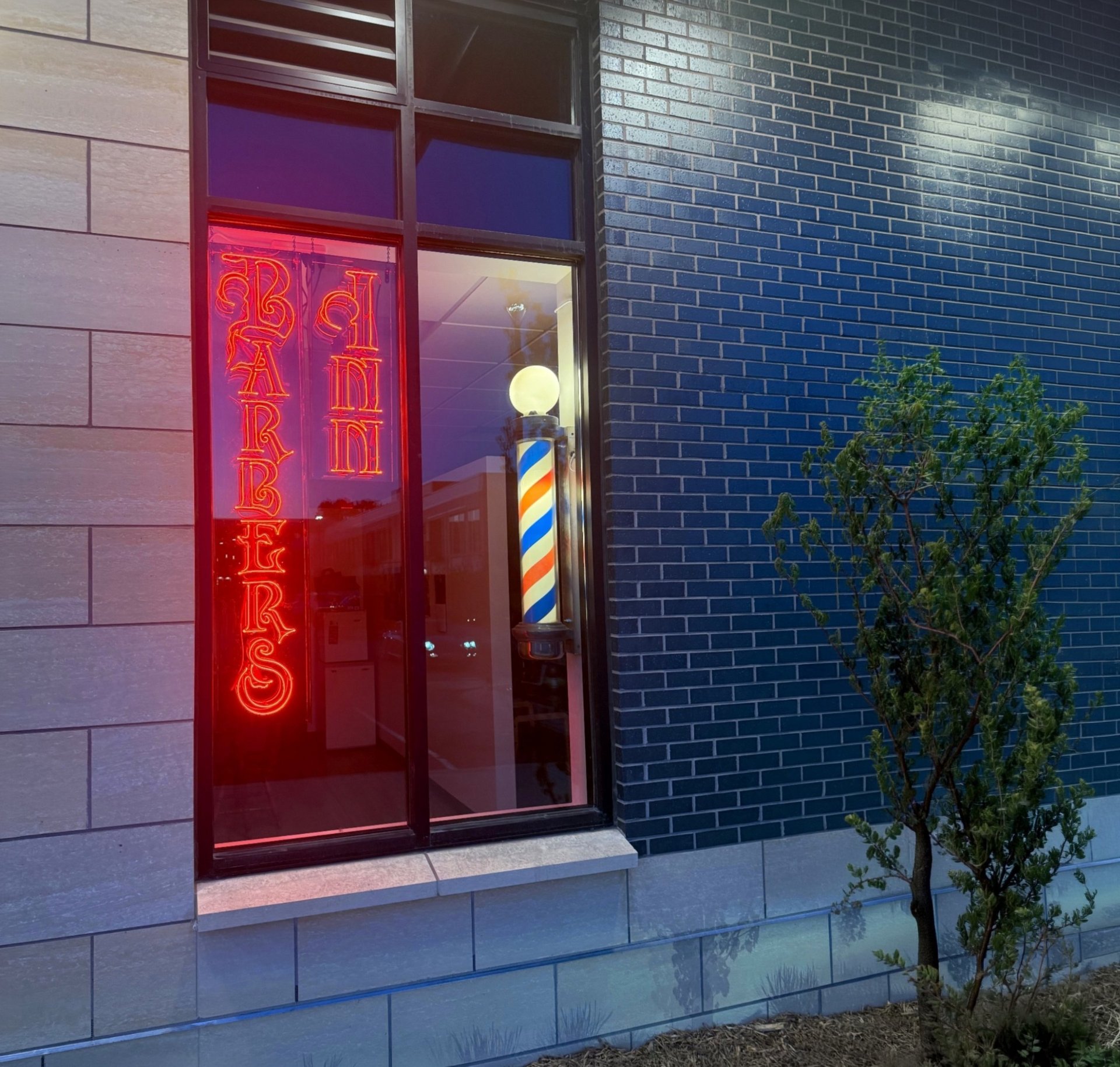 Storefront with red neon barber shop sign on window, featuring red door and blue brick wall beside it